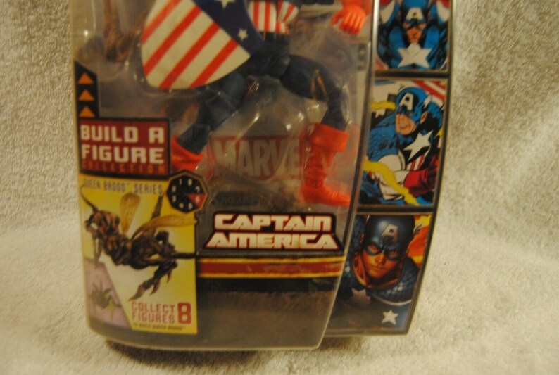 May include: Captain America action figure in packaging. The figure is wearing a blue and red costume with a shield. The packaging includes the text "Captain America", "Build a Figure Collection", and "Marvel".