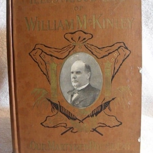 May include: A vintage book titled "Illustrious Life of William McKinley." The cover is a warm brown with gold lettering and a portrait of McKinley. The book also features decorative elements and the text "Our Martyred President."