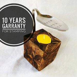 May include: A wooden candle holder with a metal spoon attached to the top. The spoon has the text "YOU TEXT HERE" engraved on it. The candle holder is brown and has a rustic look. A yellow candle is burning inside the holder.