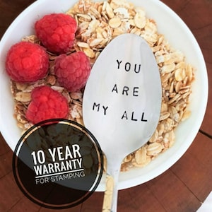 Puede incluir: Una cuchara de plata con las palabras "You are my all" estampadas en ella. La cuchara está en un tazón de cereal con frambuesas. Un círculo negro con texto blanco dice "10 años de garantía para el estampado".