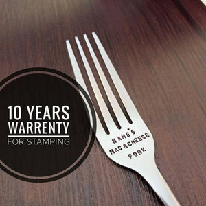Puede incluir: Un tenedor de plata con el texto "NAME'S MAC & CHEESE FORK" grabado en el mango. Un círculo con el texto "10 YEARS WARRENTY FOR STAMPING" está en el fondo.