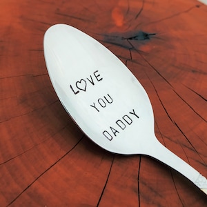 May include: A silver spoon with the words "LOVE YOU DADDY" engraved on it. The spoon is on a wooden surface.