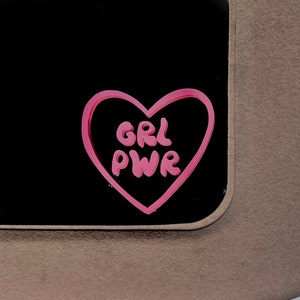 以下が含まれることがあります： 黒い背景に「GRL PWR」という文字が書かれたピンクのハート型のステッカー