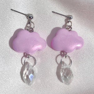 May include: A pair of earrings featuring a light pink cloud charm with a clear crystal raindrop dangling below. The earrings have silver hooks.