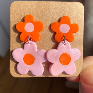 Könnte beinhalten: Ein Paar Ohrringe mit zwei übereinanderliegenden Blumenanhängern. Die obere Blume ist orange mit einem rosa Zentrum, die untere Blume ist rosa mit einem orangefarbenen Zentrum.