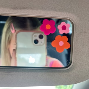May include: A car visor mirror with three flower-shaped stickers in shades of pink and orange. The mirror reflects a person holding a phone and wearing a pink hair clip. The visor is gray.
