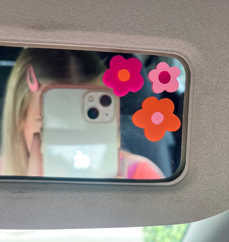 May include: A car visor mirror with three flower-shaped stickers in shades of pink and orange. The mirror reflects a person holding a phone and wearing a pink hair clip. The visor is gray.
