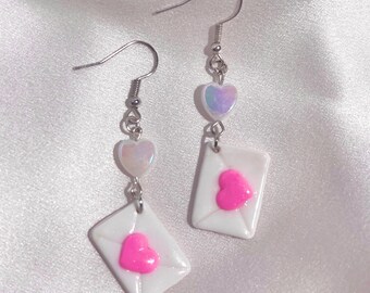 May include: A pair of dangle earrings featuring white envelope charms with bright pink heart accents. Each earring has a small iridescent heart and a silver-tone fish hook. The earrings are set against a soft, white background.