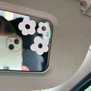 May include: Three white daisy-shaped car mirror stickers are affixed to a car visor mirror. The stickers vary in size and have a circular cutout in the center. The mirror reflects a smartphone.
