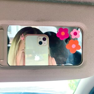 May include: A car visor mirror with three flower stickers in shades of pink and orange. The mirror reflects a person holding a phone, with pink hair clips visible. The phone has a clear case.