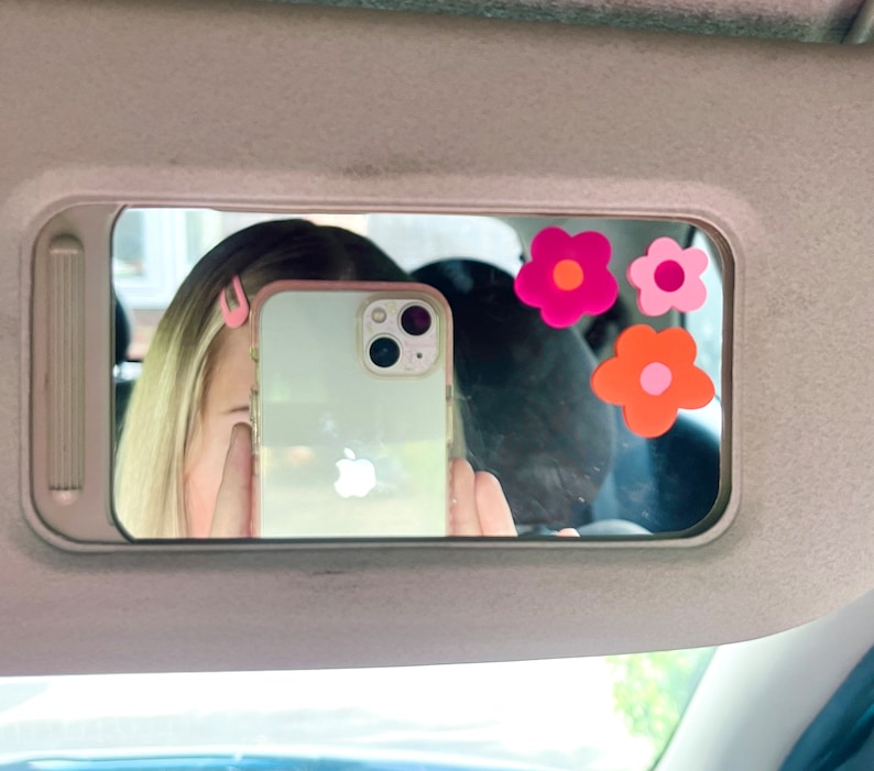 May include: A car visor mirror with three flower stickers in shades of pink and orange. The mirror reflects a person holding a phone, with pink hair clips visible. The phone has a clear case.