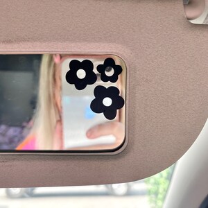 May include: Three black flower stickers with white centers are affixed to a car visor mirror. The visor is a light tan color. The mirror reflects a person holding a phone.