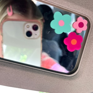 May include: A car visor with three flower-shaped decorations in shades of teal, pink, and magenta. The flowers have contrasting center circles in pink and orange. The visor is gray, and the image is reflected in a mirror.