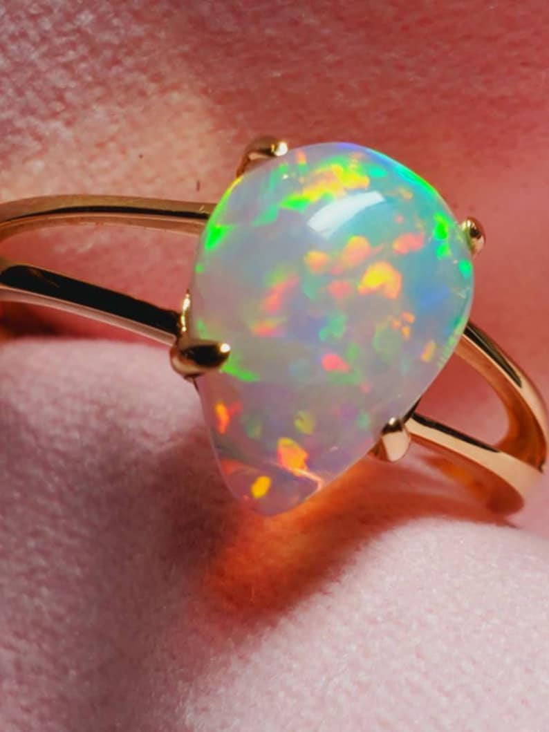 Op de afbeelding: Een gouden ring met een peervormige opaal edelsteen. De opaal heeft een witte basis met flitsen van groen, geel, oranje en rood.