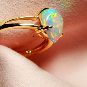 Op de afbeelding: Een gouden ring met een peervormige opaal edelsteen. De opaal heeft een witte basis met iriserende flitsen van groen, blauw en roze.