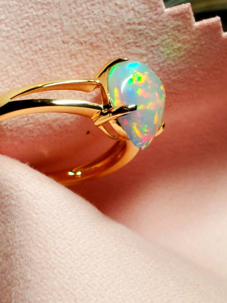 Op de afbeelding: Een gouden ring met een peervormige opaal edelsteen. De opaal heeft een witte basis met iriserende flitsen van groen, blauw en roze.