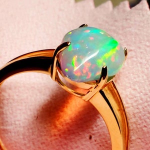 Op de afbeelding: Een gouden ring met een grote, ovale opaal. De opaal is iriserend met groene, blauwe en roze schittering.