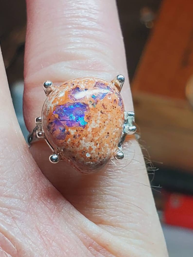 May include: A silver ring with a large, orange opal gemstone set in a prong setting. The opal has a unique pattern of purple, blue, and white.