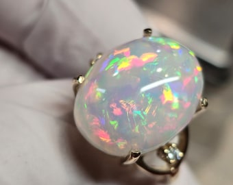 May include: A white opal ring with a gold setting. The opal has a rainbow of colors, including red, orange, yellow, green, blue, and purple.