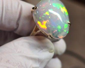 Op de afbeelding: Een gouden ring met een grote ovale opaal. De opaal is iriserend met groene, blauwe en rode schittering.