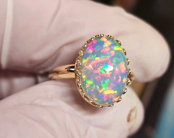 May include: A gold ring featuring a large, oval opal gemstone. The opal displays a vibrant play of color with flashes of blue, green, pink, and yellow. The ring has a detailed, ornate setting around the opal.
