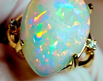 Op de afbeelding: Een gouden ring met een grote ovale opaal. De opaal heeft een witte basis met iriserende flitsen van groen, blauw en roze.