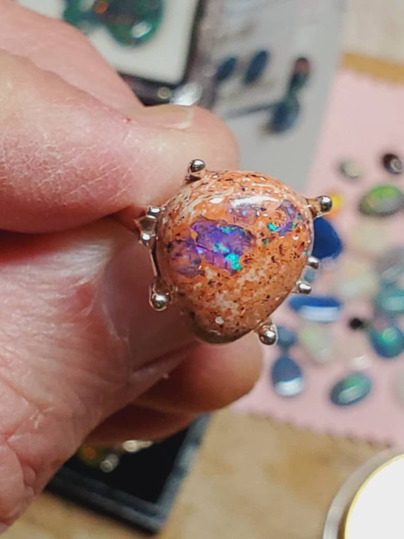 May include: A silver ring with a teardrop-shaped opal stone. The opal is a mix of orange, brown, and purple with iridescent flashes.