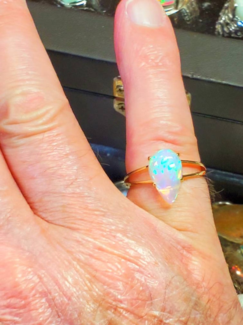 May include: A gold ring with a teardrop-shaped opal gemstone. The opal is iridescent with shades of blue, green, and white.