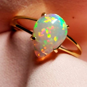 Op de afbeelding: Een gouden ring met een druppelvormige opaal-edelsteen. De opaal is wit met iriserende flitsen van groen, blauw en roze.