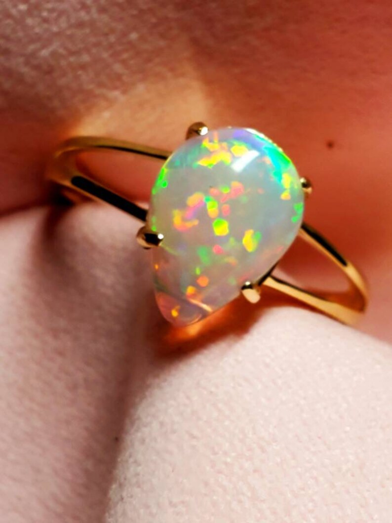 Op de afbeelding: Een gouden ring met een druppelvormige opaal-edelsteen. De opaal is wit met iriserende flitsen van groen, blauw en roze.