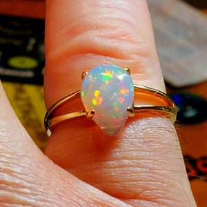 Op de afbeelding: Een gouden ring met een peervormige opaal edelsteen. De opaal is wit met iriserende flitsen van groen, blauw en rood.