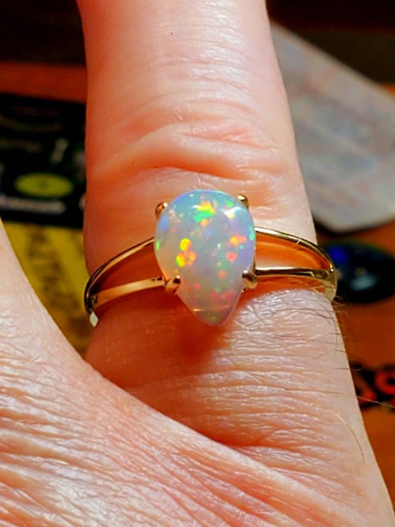 Op de afbeelding: Een gouden ring met een peervormige opaal edelsteen. De opaal is wit met iriserende flitsen van groen, blauw en rood.