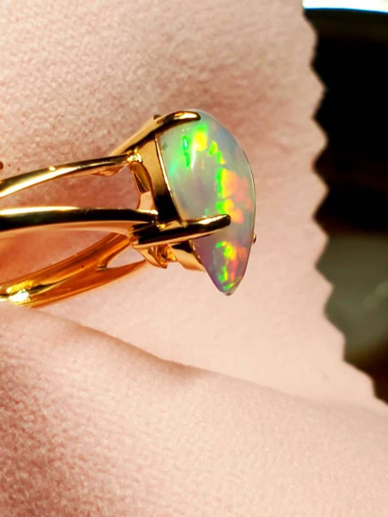 Op de afbeelding: Een gouden ring met een grote, iriserende opaal-edelsteen. De opaal heeft een witte basis met flitsen van groen, geel en oranje.