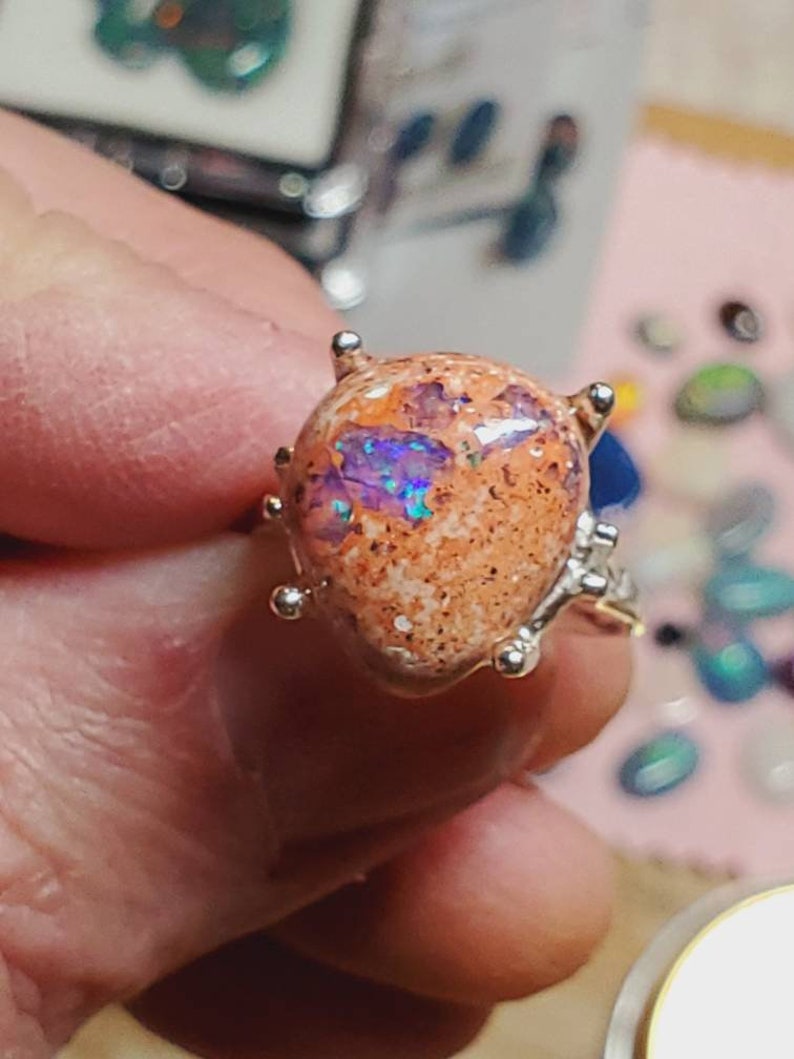 May include: A silver ring with a large, oval-shaped opal gemstone. The opal is a mix of orange, white, and purple colors with a sparkly, iridescent finish.