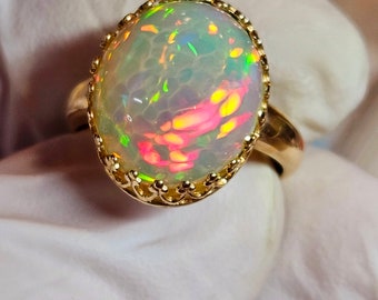 May include: A gold ring with a large oval opal gemstone. The opal has a white base with flashes of green, yellow, and pink.