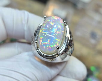 May include: A large, oval-shaped opal gemstone ring. The opal has a milky white base with iridescent flashes of color. The ring is set in a silver-colored metal band with a detailed design on the sides.
