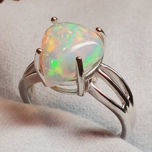 Pode incluir: Um anel de prata com uma grande pedra de opala oval. A opala tem uma base branca com flashes de verde, azul e laranja.