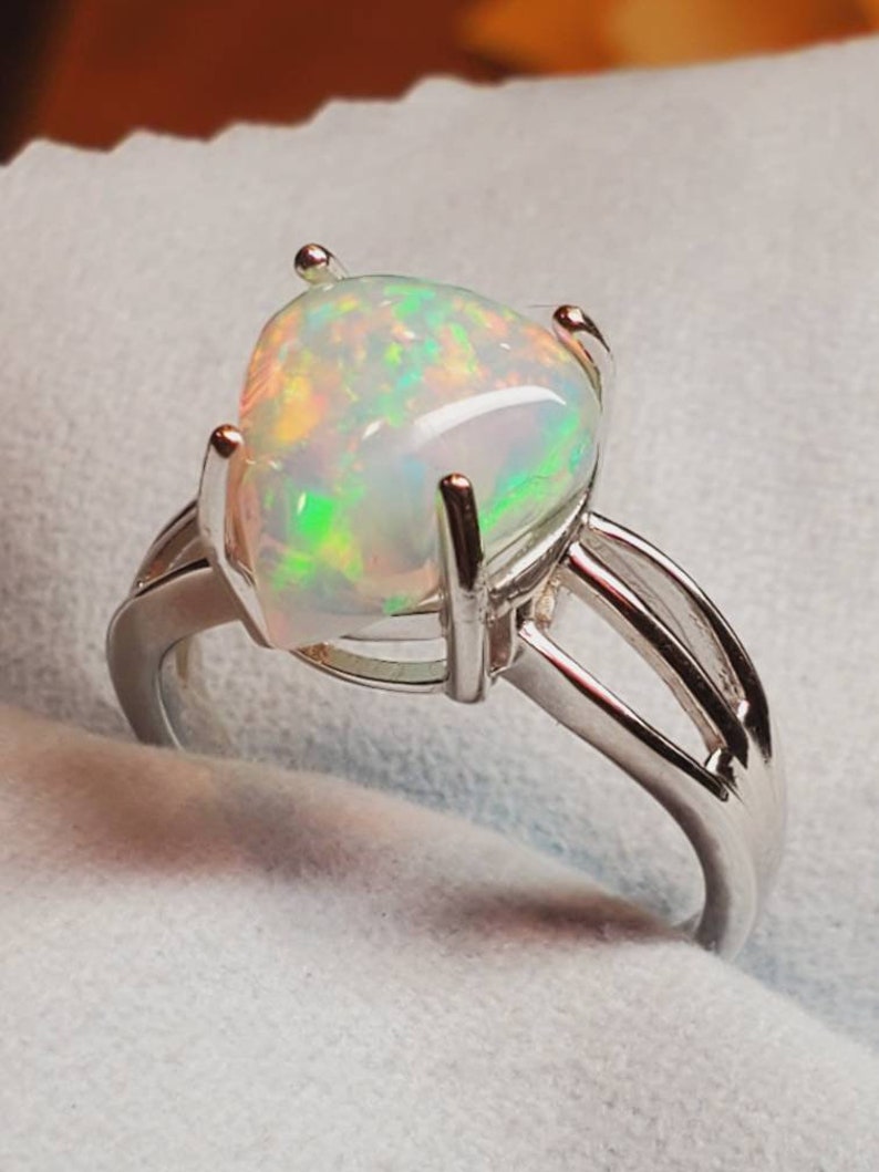 Pode incluir: Um anel de prata com uma grande pedra de opala oval. A opala tem uma base branca com flashes de verde, azul e laranja.