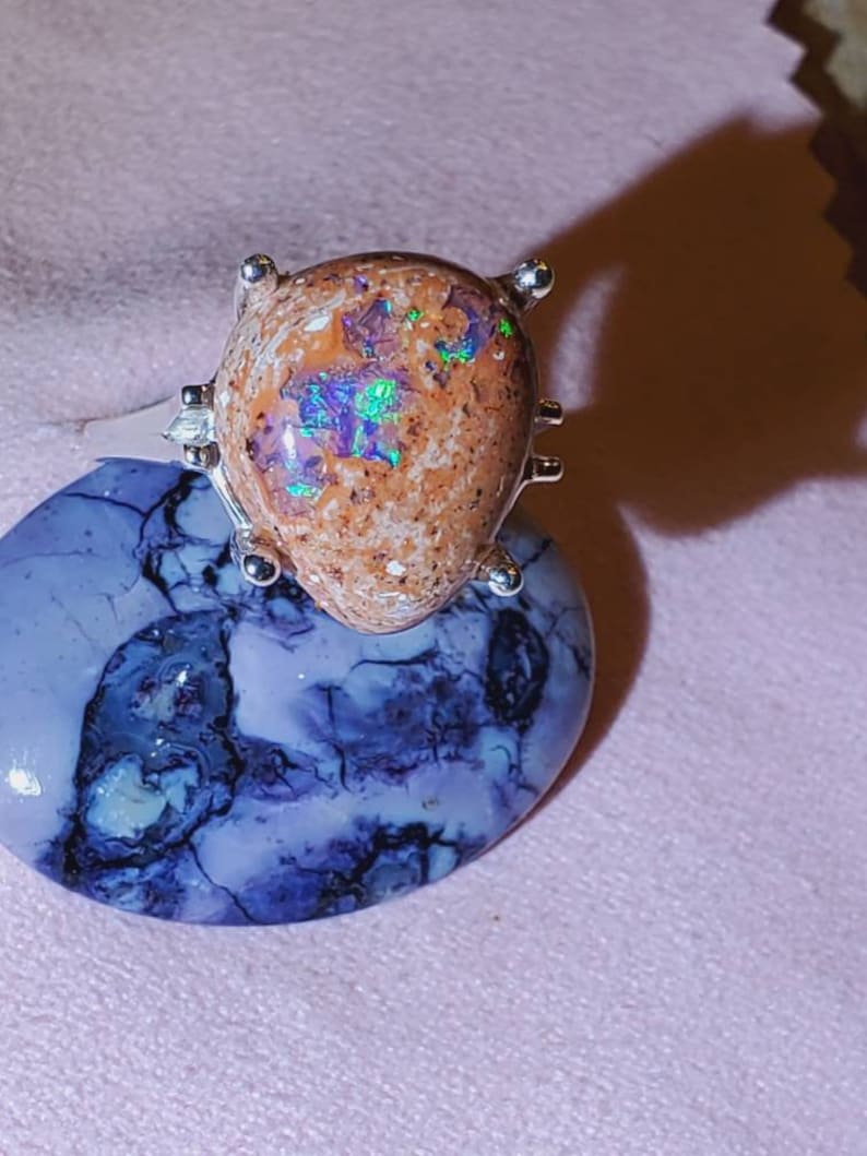 May include: A silver ring with a brown opal gemstone set in a prong setting. The opal has iridescent flashes of green, blue, and purple.