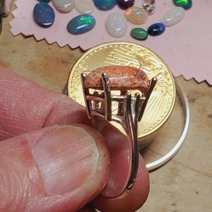 May include: A silver ring with a large, oval-shaped, pink and white opal gemstone set in a prong setting. The ring is on a finger and a gold coin is in the background.