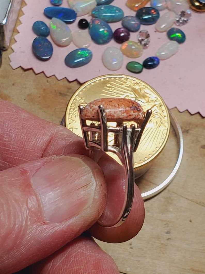 May include: A silver ring with a large, oval-shaped, pink and white opal gemstone set in a prong setting. The ring is on a finger and a gold coin is in the background.