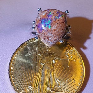 May include: A silver ring with a brown and iridescent opal gemstone. The ring is set on a gold coin with the image of Lady Liberty and the year 1993.