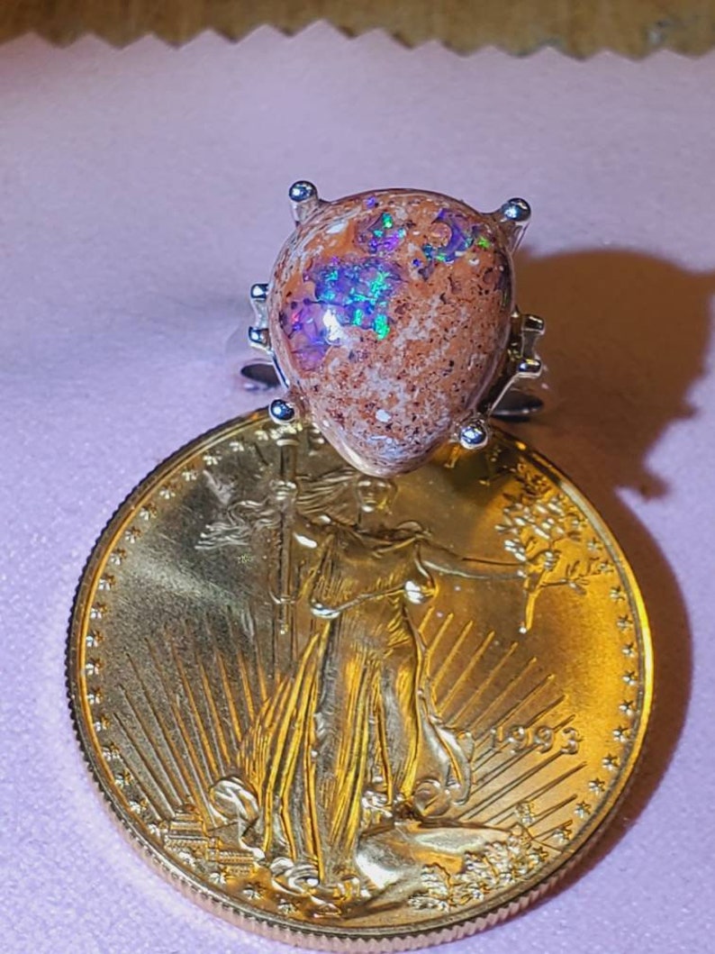 May include: A silver ring with a brown and iridescent opal gemstone. The ring is set on a gold coin with the image of Lady Liberty and the year 1993.
