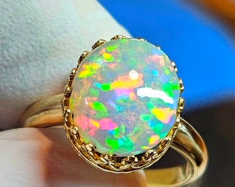 May include: A gold ring featuring a large, oval opal gemstone. The opal displays a vibrant play-of-color with flashes of green, pink, and yellow. The ring has a detailed, ornate gold setting, showcasing the gemstone.