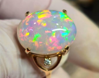 May include: A gold ring featuring a large, oval-shaped cabochon opal with vibrant flashes of pink, green, and blue iridescence. The setting includes a small, round, clear faceted gemstone positioned directly below the central opal stone.