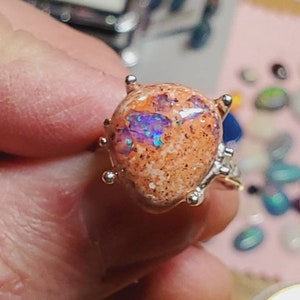 May include: A silver ring with a large, oval-shaped opal gemstone. The opal is a mix of orange, white, and purple colors with a sparkly, iridescent finish.