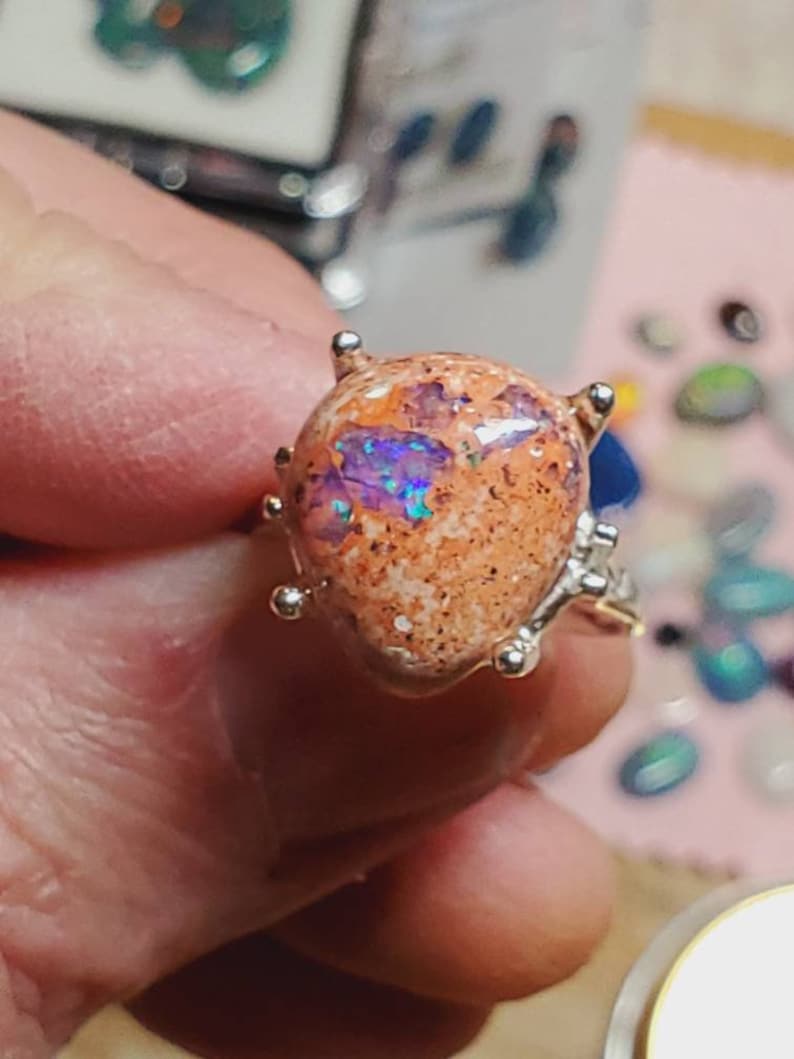 May include: A silver ring with a large, oval-shaped opal gemstone. The opal is a mix of orange, white, and purple colors with a sparkly, iridescent finish.