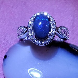 Puede incluir: Un anillo de plata con una piedra preciosa de ópalo ovalada de color azul oscuro rodeada por un halo de piedras preciosas más pequeñas y transparentes.