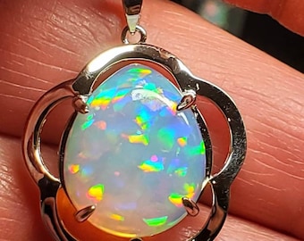 May include: A pendant necklace featuring a large, oval opal gemstone with iridescent flashes of color. The opal is set in a gold-colored, flower-shaped frame and hangs from a delicate chain. The gemstone displays a range of colors, including blue, green, and yellow.