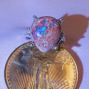 May include: A silver ring with a large, pear-shaped opal gemstone. The opal is a mix of brown, white, and green with iridescent flashes of color. The ring is sitting on top of a gold American Eagle coin.